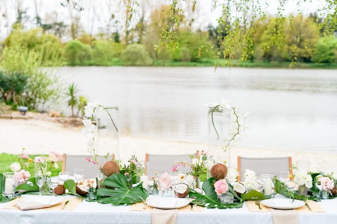 Table élégamment dressée au bord d'un lac, décorée de fleurs et de verdure.