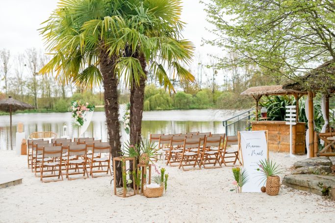Cérémonie en plein air avec chaises en bois face à un plan d'eau et des palmiers.