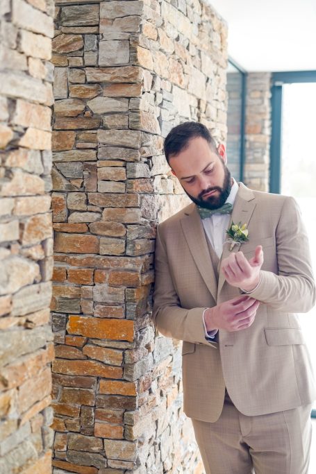 Homme en costume clair ajustant sa montre, près d'un mur en pierre.