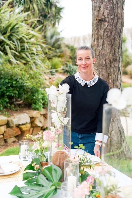 Femme souriante en pull noir, entourée de décorations florales en extérieur.